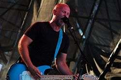 Bob Mould—a middle-aged Caucasian man with a shaved head and short facial hair wearing a black t-shirt—plays a blue guitar and sings into a microphone