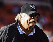 Candid head-and-shoulders photograph of Wylie sporting a bushy blond-grey mustache and wearing a black baseball cap bearing an Oakland Raiders logo