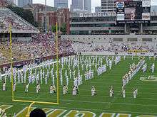 A football stadium with a marching band in white uniforms on the field, with the goal post in the foreground and various buildings in the background