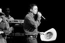 A black and white photo of Pulido wearing a dress shirt tucked in jeans with his cowboy hat on one hand and a microphone on the other. A Keyboardist and his instrument can be seen in the foreground of the photo.
