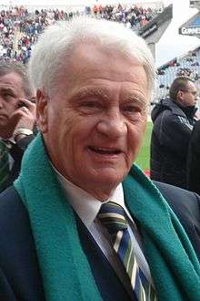 An elderly man with grey hair smiling at the camera. He is in a football stadium and is wearing a black suit with a multi-coloured tie and a green scarf.