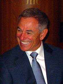A gray-haired man in a dark blue suit and white shirt smiles.