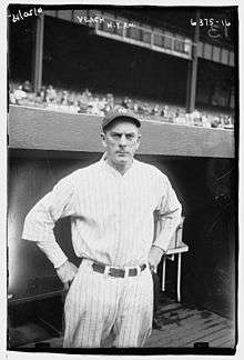 A man in a white baseball uniform stands in front of a dugout with his hands on his hips.