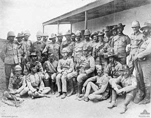 Group of soldiers gathered in a semicircle for an informal portrait. They are wearing a variety of different headgear including pith helmets, garrison caps and slouch hats. An unsmiling Chauvel wears a bandolier and slouch hat, and holds a rifle.