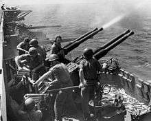 The gun crew operating these guns; four men handle ammunition while another yells over the blast from the guns. Spent shell casings litter the deck below.