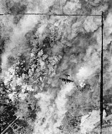 An overhead view of a bomber aircraft, flying over a pall of smoke