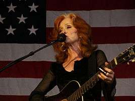 A woman with red hair wearing black clothing and hoop earrings playing the guitar and singing into a microphone. The American flag appears in the background.