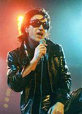 Bono with black hair, black sunglasses, and a black leather attire speaking into a microphone.