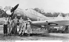 Six men in front of a single-engined military monoplane parked on a jungle airfield