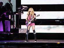 Image of a blond female singing to a microphone and playing a purple electric guitar. She's wearing a pink ensemble and knee-high stockings. To her right there are 2 male musicians.