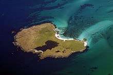 An island seen from above sits in a dark green water and sandy shallows. The island has some sandy beaches and contains a body of water that makes up about a sixth of its area.