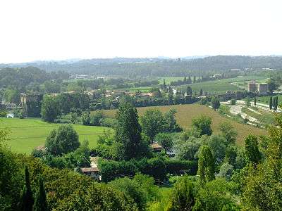 Photo overlooks the countryside on the west side of the Mincio near Borghetto. The old bridge is at the left and the Mincio loops from there to the right foreground amid trees.