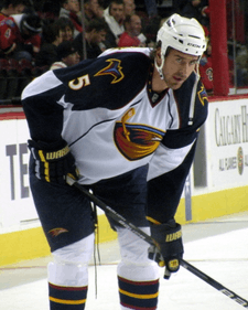 A Caucasian hockey player. He wears a white jersey with dark blue shoulders with a stylized brown thrasher holding a hockey stick logo with the number 5 on his sleeve. He is bent over at the waist leaning on his stick.