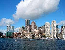Skyline of a waterfront city. Several skyscrapers are visible in the distance, the tallest being a building with rounded edges and a red facade at the center of the image. To the extreme right is a white clock tower that appears shorter than most of the other tall buildings; two concrete, Brutalist-era apartment buildings are adjacent to the clock tower.