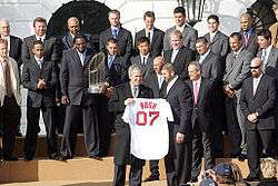 Several men in suits stand behind a man holding a white baseball jersey which reads "BUSH 07" on it.