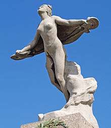 A photograph from ground level looking up towards a memorial statue of a flying woman on top of a pillar. She is nude, her arms are outstretched with a drape or wing behind her, and her face is lifted towards the sky.