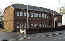 Three-quarter view of a two-storey brown brick building with a shallow, grey, partly hipped roof.  The two storeys are separated by a thin band of projecting bricks.  Each floor has three sets of four rectangular windows.  To the right, a projecting section includes a round-headed entrance door, the words "BOWLS ENGLAND" on a white background, and a red and blue logo consisting of a heraldic lion and a crown.