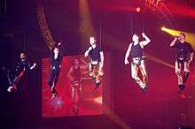 Colour photograph of all five members of Boyzone performing in concert in 2009. They are suspended from the ceiling by wires.