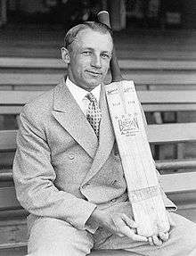 Cricketer Donald Bradman sits on a bench in a sports stadium and displays a cricket bat with his brand name on it. He wears a 1940s-style double-breasted suit and has his hair parted and slicked back with haircream. He is smiling and posing rather awkwardly.