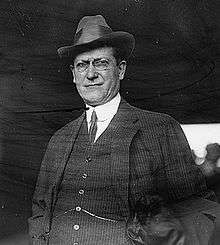 Black and white photo of a white man in a three-piece suit with glasses and a hat.