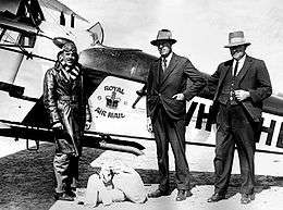 Three men, two in suits and hats, the other in flying gear, standing in front of biplane bearing the legend "Royal Air Mail"