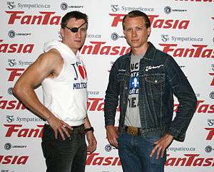 Two men in their mid-20s stand with their hands at their hips and facing the camera. The man on the left, Jason Trost, has short brown hair and wears an eyepatch, a sleeveless hooded sweatshirt, and black jeans. The man on the right, Brandon Trost, also has short brown hair, wears a denim jacket over a gray t-shirt with an unreadable graphic, and blue jeans.