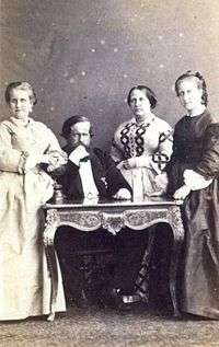 A photograph showing a bearded man seated at a table with an older woman with dark hair standing immediately to his left, and two younger women in long, mid-Victorian dresses standing one at the left side of the table and one at the right side of the table