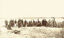 Photograph of a line of uniformed soldiers ranged along a river bank in the foreground with boats on the river in the background