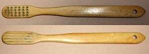 Front and back view of a light brown toothbrush. The back has four grooves running lengthwise along the head. The front has five rows of holes at the head and an inscription on the handle