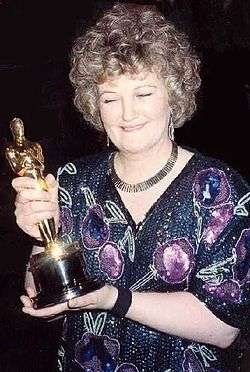 A picture of a curly-haired woman wearing a dress with various purple and green prints. She is holding a golden statuette.