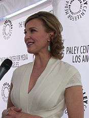 A blond woman shown smiling as she is being interviewed with a microphone pointed towards her. She is wearing a white dress with turquoise earrings, and is standing behind a backdrop with the words "THE PALEY CENTER FOR MEDIA" followed by the words "LOS ANGELES".