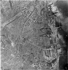 Vertical aerial photograph taken during a daylight attack on German warships docked at Brest, France. Two Handley Page Halifaxes of No. 35 Squadron RAF (upper right) fly over the naval dockyard, towards the dry docks in which the battlecruisers Scharnhorst and Gneisenau are berthed (top right) and over which a smoke screen is rapidly spreading. At middle right, a stick of bombs can be seen to have exploded inland from their intended target, Prinz Eugen, moored by the quayside. 47 aircraft from Nos. 3, 4 and 5 Groups took part in the operation, claiming accurate bombing on their targets, for the loss of 6 aircraft