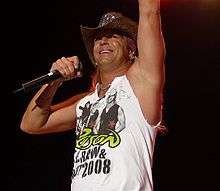 A man wearing a cowboy hat and a sleeveless concert T-shirt for the band Poison; one arm is raised while the other holds a microphone.