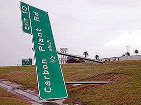 A sign on its side, blown down by high winds, for Exit 10 that reads "Carbon Plant Rd ½ mile". A building and another large, green exit sign can be seen in the background.