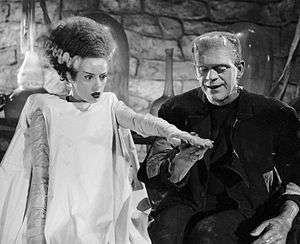 The Bride of Frankenstein has black hair with a white streak running through it, is dressed in a white gown, and has a blank expression. She is standing on the left with her left hand elevated. On the right is Frankenstein's monster, standing on the right and smiling. His right hand is below hers. The background includes walls made of stone.