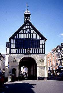 A photograph of a building on a clear summer's day, with a white stone base and a black and white timbered first floor. An archway leads through the middle of the building, apparently located in the middle of a small town.