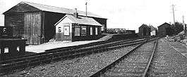 small wooden hut with a large wooden shed behind it. In front of the hut is a platform at two different heights, and in front of the platform are two sets of railway tracks. The tracks lead into a further pair of wooden sheds.