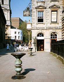 Two ornate metal pillars with large dishes on top in a paved street, with an eighteenth-century stone building behind, upon which can be seen the words "Tea Blenders Estabklishec 177-". People sitting at café-style tables outside. On the right are iron railings.