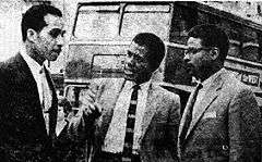 Three Afro-Caribbean men in conversation; behind them can be seen the upper part of a double-decker bus.