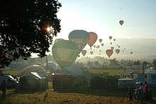 A large number of hot air balloons taking off from a field which is surrounded by tents and stalls. The sun is low in the sky and balloons can be seen flying into the distance.