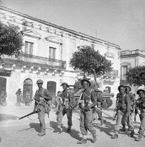 File of soldiers walking through a town with a tank in the background