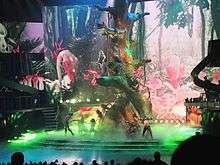 A blond woman female performer wearing a black outfit on top of a giant tree, while other people look at her.