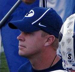 A white man wearing a blue hat with the Brigham Young University Logo partially visible addresses his team (not pictured), with part of the helmet of a player is visible in the foreground.