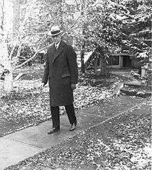 A man in a hat and overcoat walks down the front walk of a house. There is snow on the ground.