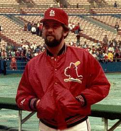 A bearded man with his hands in his jacket pocets, wearing a red baseball cap and red jacket with a bird logo on the left chest, standing with a field and stands in the background.