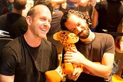 A man with short brown hair, sitting next to a man with curly black hair hugging a plush giraffe, both smiling at something to the right of the camera.