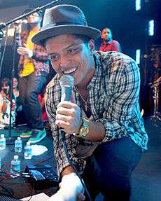 Bruno Mars down on one knee at the front of a stage, singing into a microphone, with someone in the audience holding his forearm. He is wearing black pants, a plaid button-up shirt, and a gray fedora.
