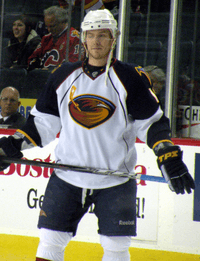 A Caucasian hockey player. He wears a white jersey with dark blue shoulders with a stylized brown thrasher holding a hockey stick for a logo. He holds his stick parallel to his waist.