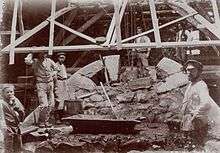 A sepia photograph showing six workers standing under a wooden scaffolding erected over a pile of stones, with a trough for mixing cement visible in the centre.