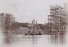 Black and white (sepia) photo showing construction of a bridge across the Neman. On both sides completed parts of the bridge surrounded by wooden scaffolding are visible. In between a wooden pyramid-shaped floating crane is being assembled.